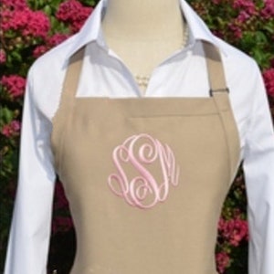 May include: A tan apron featuring a light pink embroidered monogram with the letters SSM in a cursive script font. The apron is worn over a white collared shirt, with the fabric covering the chest area and featuring adjustable straps.