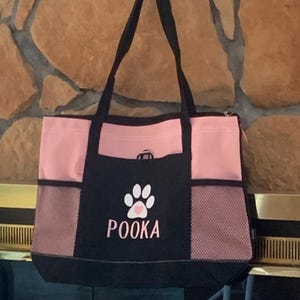 May include: A pink and black tote bag with a paw print and the name "POOKA" embroidered on the front. The bag has a mesh pocket on the side and a zipped pocket on the top.