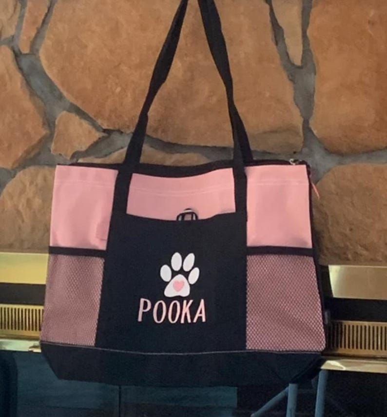 May include: A pink and black tote bag with a paw print and the name "POOKA" embroidered on the front. The bag has a mesh pocket on the side and a zipped pocket on the top.