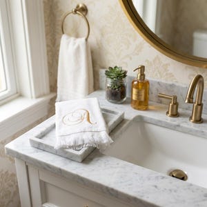 May include: A white hand towel with a gold embroidered letter A and fringed edges, resting on a rectangular white marble tray next to a marble bathroom vanity with a gold faucet, a glass soap dispenser, and a small potted succulent plant.