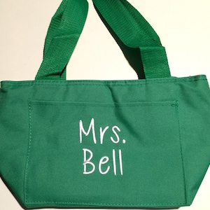 May include: A bright green tote bag featuring two matching green fabric handles and a front pocket. The text "Mrs. Bell" is embroidered in white thread on the front pocket. The bag is constructed from a solid green fabric with visible stitching.
