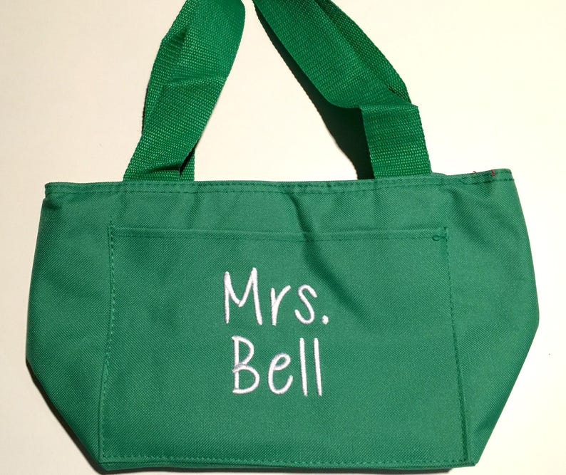 May include: A bright green tote bag featuring two matching green fabric handles and a front pocket. The text "Mrs. Bell" is embroidered in white thread on the front pocket. The bag is constructed from a solid green fabric with visible stitching.