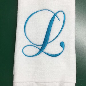 May include: A white rectangular hand towel featuring a large, embroidered cursive letter L in a bright turquoise blue thread. The towel has a textured terry cloth surface and a decorative fringe trim along the bottom edge.