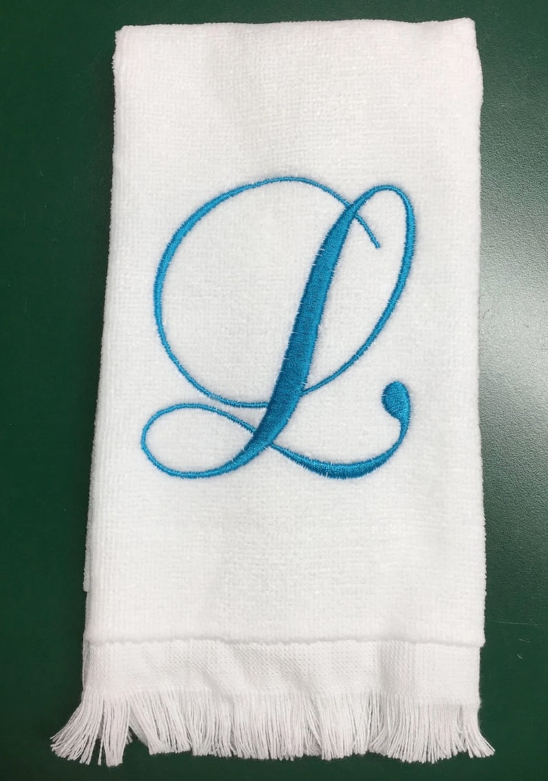 May include: A white rectangular hand towel featuring a large, embroidered cursive letter L in a bright turquoise blue thread. The towel has a textured terry cloth surface and a decorative fringe trim along the bottom edge.