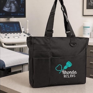 May include: A black tote bag with two shoulder straps, multiple exterior pockets, and a teal graphic of a heart-shaped ultrasound probe. The text "Rhonda RCS, RVS" is printed in white below the graphic. The bag features a zipper closure.