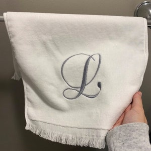 May include: A white hand towel featuring a centered, embroidered cursive letter L in a soft grey thread. The towel has a smooth texture and is finished with a delicate fringe along the bottom edge, providing a clean and simple aesthetic for bathroom decor.