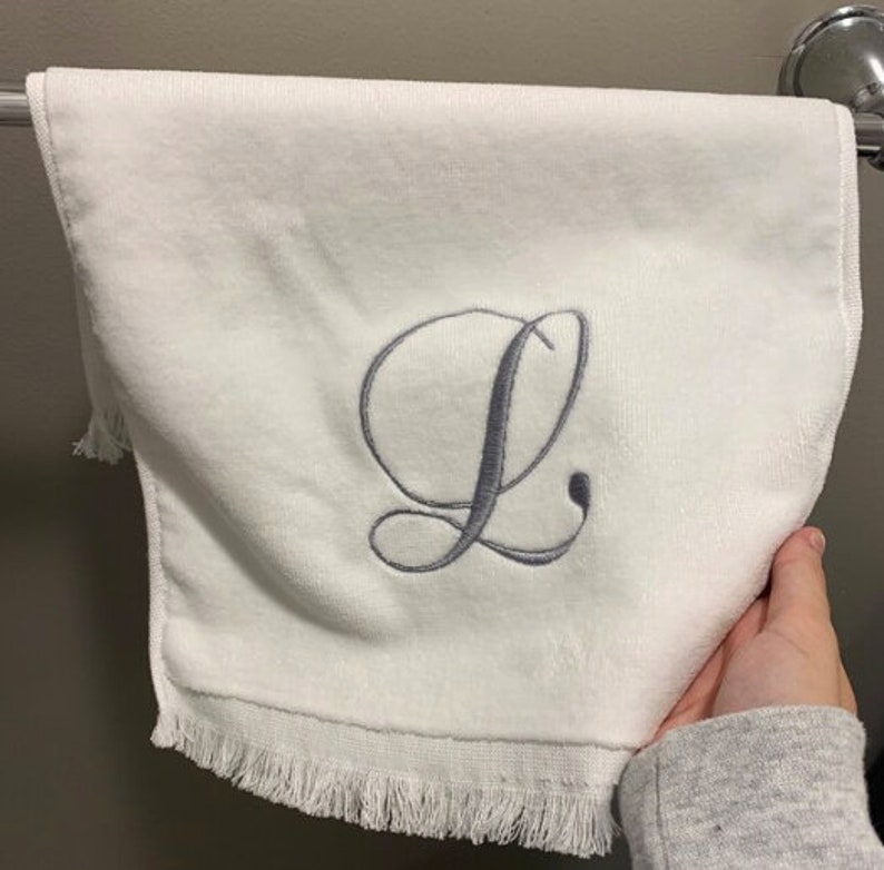 May include: A white hand towel featuring a centered, embroidered cursive letter L in a soft grey thread. The towel has a smooth texture and is finished with a delicate fringe along the bottom edge, providing a clean and simple aesthetic for bathroom decor.
