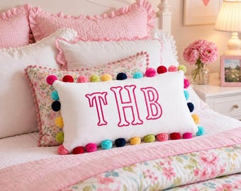 May include: A rectangular white throw pillow featuring the embroidered pink monogram letters THB in the center. The pillow is decorated with a border of small, multicolored pom-poms in shades of pink, blue, yellow, and green.