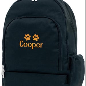 May include: Black backpack with orange paw prints and the name "Cooper" embroidered on the front. The backpack has a top handle, two zippered compartments, and a side pocket. The zippers are silver.