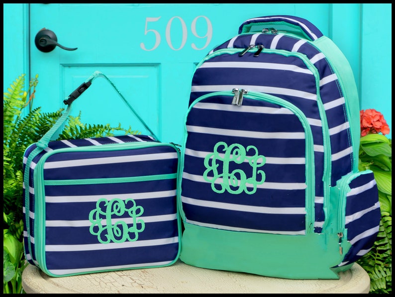 May include: A navy blue and white striped backpack and matching lunch bag set. Both items feature a bright mint green trim and a decorative mint green monogrammed letter C in a swirling script font on the front. The backpack has silver zippers.