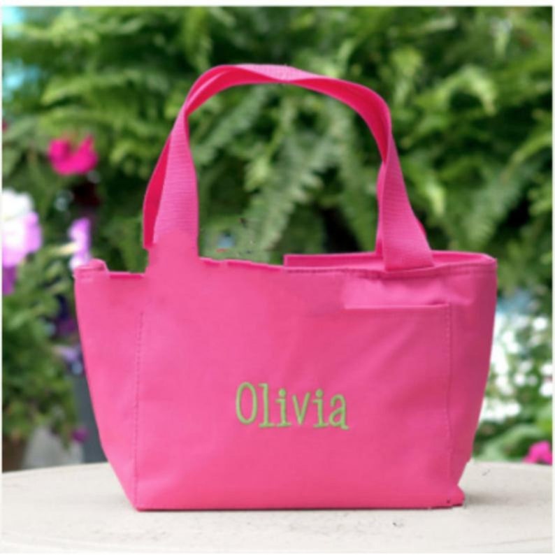 May include: A bright pink tote bag featuring two sturdy handles and a single exterior pocket. The name Olivia is embroidered in light green thread across the center of the front panel, providing a clear and personalized detail on the bag.