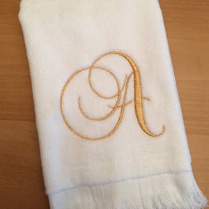 May include: A white rectangular hand towel featuring a decorative gold embroidered letter A in a cursive script font. The towel has a soft, plush texture on the main body and a fringe trim along the bottom edge, creating a clean and simple look.