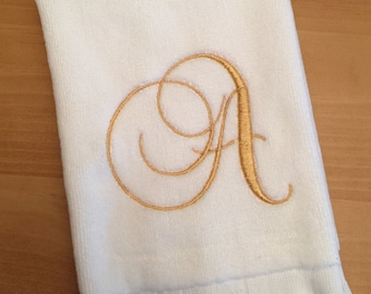 May include: A white rectangular hand towel featuring a decorative gold embroidered letter A in a cursive script font. The towel has a soft, plush texture on the main body and a fringe trim along the bottom edge, creating a clean and simple look.