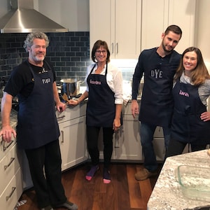 Puede incluir: Cuatro personas en una cocina con delantales azul marino. Los delantales est&aacute;n bordados con los nombres "Papa", "Grandma", "DYE" y "Sarah" en hilo rosa. La cocina tiene armarios blancos, una campana extractora de acero inoxidable y una pared salpicada de azulejos azules oscuros.