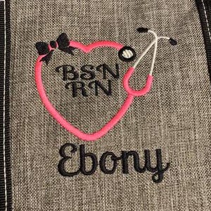 May include: Gray fabric with a pink heart-shaped stethoscope design. The design includes the letters "BSN RN", a black bow, and the name "Ebony" in black script. The stethoscope is white and pink.