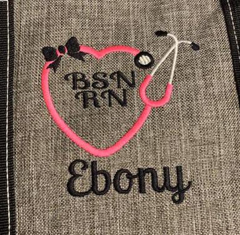 May include: Gray fabric with a pink heart-shaped stethoscope design. The design includes the letters "BSN RN", a black bow, and the name "Ebony" in black script. The stethoscope is white and pink.