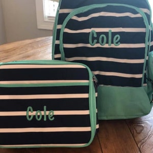May include: A navy blue and white striped backpack and matching lunch bag set. Both items feature the name Cole embroidered in light teal thread. The backpack has a large main compartment and a front pocket, while the lunch bag is rectangular.