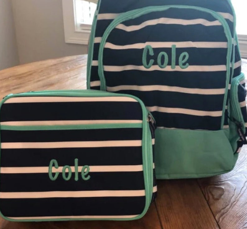 May include: A navy blue and white striped backpack and matching lunch bag set. Both items feature the name Cole embroidered in light teal thread. The backpack has a large main compartment and a front pocket, while the lunch bag is rectangular.