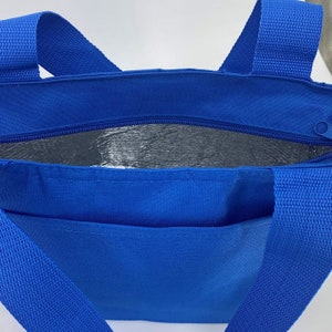 May include: A bright blue insulated tote bag featuring two sturdy woven handles and a top zipper closure. The interior is lined with a reflective silver thermal material designed for temperature control, and the exterior includes a front pocket.