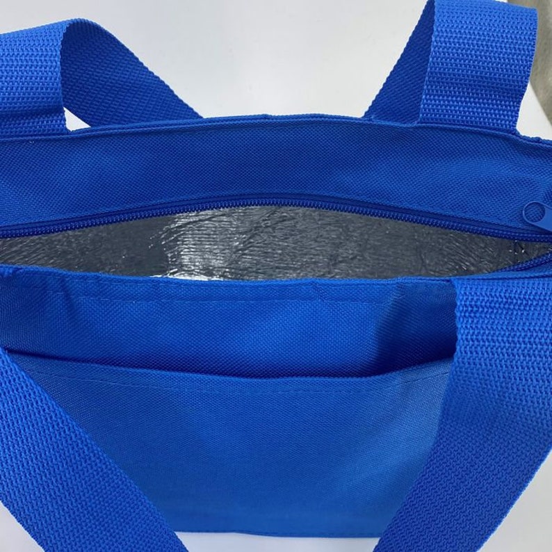 May include: A bright blue insulated tote bag featuring two sturdy woven handles and a top zipper closure. The interior is lined with a reflective silver thermal material designed for temperature control, and the exterior includes a front pocket.