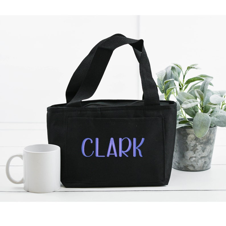 May include: A black rectangular lunch bag with two black fabric handles and a zipper closure. The front of the bag features the name CLARK embroidered in light purple capital letters. The bag is positioned next to a plain white ceramic mug.