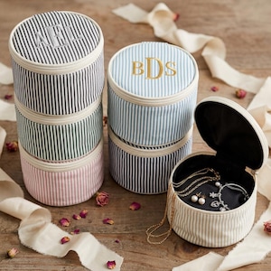 May include: A collection of round, zippered jewelry cases featuring vertical pinstripes in navy, green, pink, light blue, and beige. Each case has a cream-colored zipper and a black interior. Some cases are embroidered with monograms like AFL or BDS.