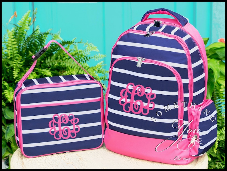May include: A navy blue and white striped backpack and matching lunch bag set with bright pink trim and zippers. Both items feature a pink embroidered monogram of the letter C on the front. The backpack includes a top handle and side pockets.