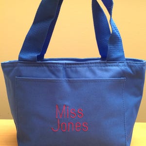 May include: A bright blue tote bag featuring two matching blue fabric handles and a front pocket. The text "Miss Jones" is embroidered in red thread on the center of the front pocket, providing a clear contrast against the blue fabric.