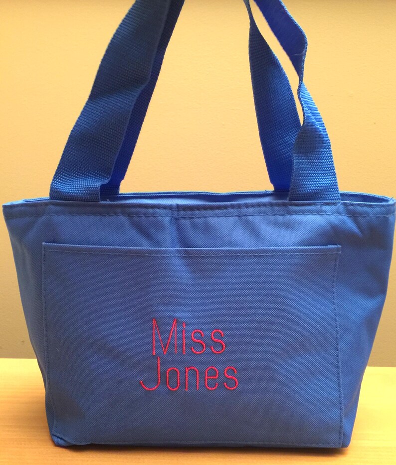 May include: A bright blue tote bag featuring two matching blue fabric handles and a front pocket. The text "Miss Jones" is embroidered in red thread on the center of the front pocket, providing a clear contrast against the blue fabric.