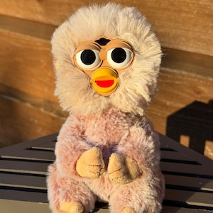 May include: A vintage Furby toy with a plush, pink body and a beige head. The toy has large, black eyes, a yellow beak, and beige feet and hands. The Furby is sitting on a dark surface against a wooden background.