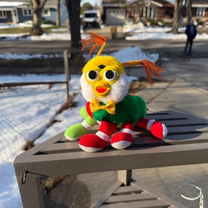 May include: A colourful plush toy with a yellow head, white fur, and large black eyes. It has a green body, red and white legs, and a yellow and green bow tie. The toy is on a grey wooden table outdoors.