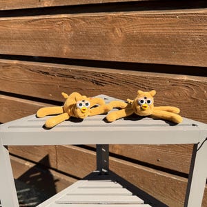 May include: Two plush, yellow toy creatures with large, round eyes and outstretched arms are displayed on a gray, triangular table. The toys have a soft, fuzzy texture and a whimsical design, suggesting a playful theme.