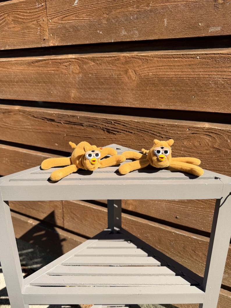 May include: Two plush, yellow toy creatures with large, round eyes and outstretched arms are displayed on a gray, triangular table. The toys have a soft, fuzzy texture and a whimsical design, suggesting a playful theme.