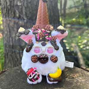 May include: A white plush toy with pink ears and large, round eyes, decorated to resemble an ice cream sundae. It features a chocolate cone, nuts, sprinkles, and fruit toppings, creating a whimsical and playful design.