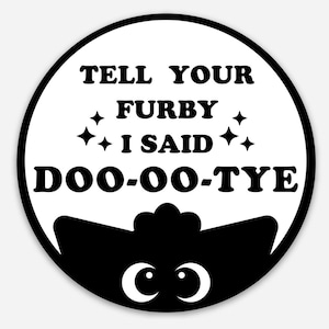 May include: A circular sticker with a black border. The white centre has the text "TELL YOUR FURBY I SAID DOO-OO-TYE" in black. A black Furby silhouette with white eyes is at the bottom of the sticker.