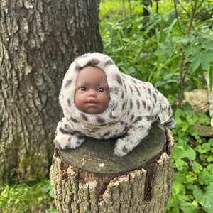 May include: A baby doll with dark skin and brown eyes is in a plush, hooded outfit with a spotted pattern. The doll is resting on a tree stump outdoors, surrounded by greenery.
