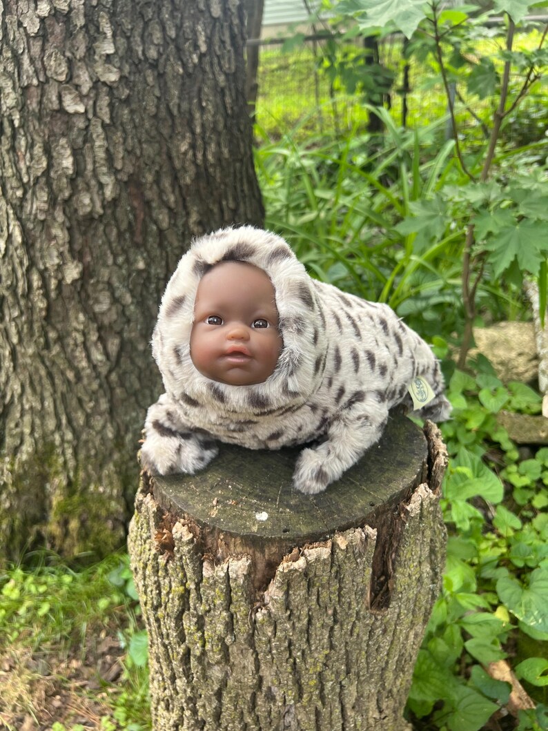 May include: A baby doll with dark skin and brown eyes is in a plush, hooded outfit with a spotted pattern. The doll is resting on a tree stump outdoors, surrounded by greenery.