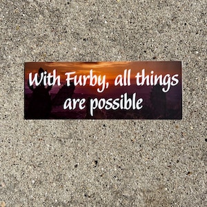 Könnte beinhalten: Ein rechteckiger Aufkleber mit dem Text "With Furby, all things are possible" in weißer Schrift. Der Hintergrund zeigt einen Sonnenuntergang mit Orange-, Lila- und Schwarztönen. Der Aufkleber liegt auf einer strukturierten grauen Oberfläche.