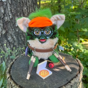Puede incluir: Un juguete de peluche verde y marrón con gafas de sol naranjas y una diadema, sosteniendo dos palos de madera y una caja de pizza. El juguete está sentado en un tocón de árbol.