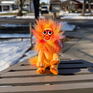 May include: A plush Furby toy in orange and yellow hues, featuring a red beak and large, round eyes. The toy has a fluffy, spiky mane and is positioned on a grey surface. A small red tag is attached.
