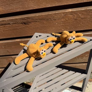 May include: Two plush, orange-yellow spider-like toys with long, flexible limbs and large, expressive eyes. The toys are lying on a gray, slatted shelf, suggesting a playful or decorative theme. The background is a wooden wall.