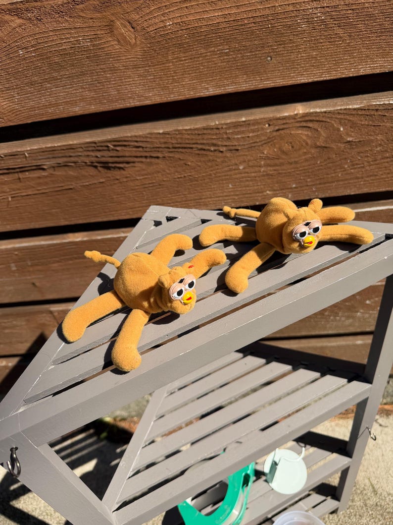 May include: Two plush, orange-yellow spider-like toys with long, flexible limbs and large, expressive eyes. The toys are lying on a gray, slatted shelf, suggesting a playful or decorative theme. The background is a wooden wall.