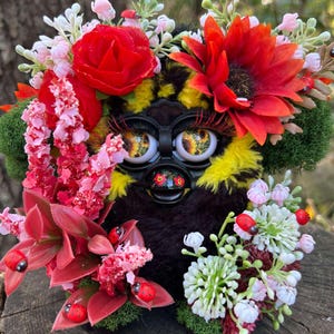 Puede incluir: Un juguete de peluche negro y amarillo con ojos grandes y una corona de flores. La corona está hecha de flores rojas, rosas y blancas con hojas verdes. También hay pequeñas mariquitas rojas esparcidas alrededor del juguete.