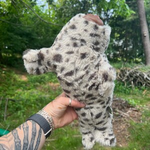 May include: A snow leopard plush toy, cream-coloured with dark brown spots. The soft toy is held against a backdrop of green foliage. The toy has a furry texture.