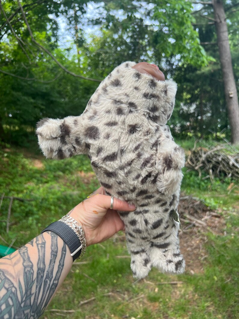 May include: A snow leopard plush toy, cream-coloured with dark brown spots. The soft toy is held against a backdrop of green foliage. The toy has a furry texture.