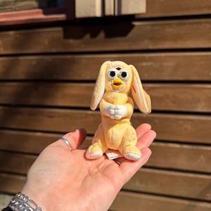 May include: A small, yellow plush toy with large eyes and long, floppy ears. The toy has a beak and is in a seated position, with its arms crossed. The background is a wooden wall.