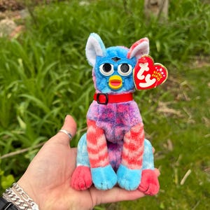 May include: A colorful plush toy with blue, pink, and red accents. The toy has large, round eyes, a yellow beak, and a red collar. A red heart-shaped tag is attached. The toy is a collectible item.