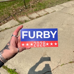Könnte beinhalten: Ein rechteckiger Aufkleber mit dem Wort "FURBY" in weißen Großbuchstaben auf blauem Hintergrund. Darunter ist die Zahl "2028" mit roten Sternen darüber und darunter dargestellt. Der Aufkleber wird vor einer Betonoberfläche gehalten.