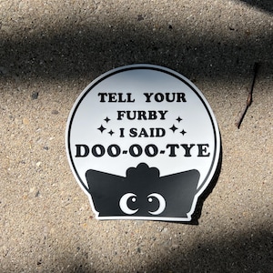 Könnte beinhalten: Rundes schwarz-weißes Schild mit dem Text "TELL YOUR FURBY I SAID DOO-OO-TYE". Das Schild hat ein cartoonartiges Design mit einer Krone und Augen am unteren Rand. Auf einer Betonoberfläche.