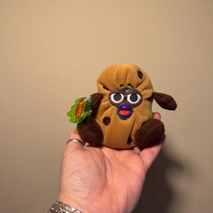May include: A plush toy shaped like a potato with a smiling face, dark brown spots, and small arms and legs. The toy has large, round eyes and a red mouth. A green tag is attached to the toy.